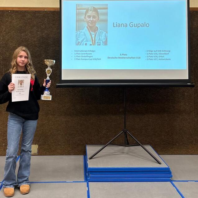 M&auml;dchen h&auml;lt eine Urkunde und einen Pokal vor einer Bildschirmpr&auml;sentation mit ihrem Namen und Foto.