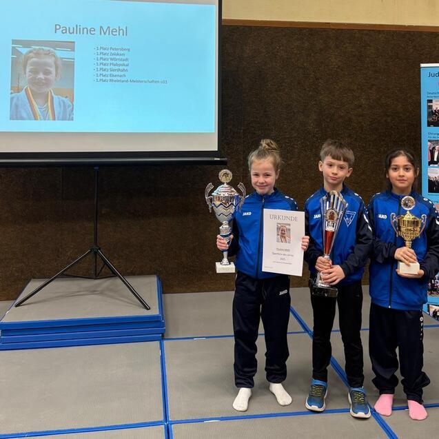 Drei Kinder stehen auf einer B&uuml;hne, halten Pokale und eine Urkunde, hinter ihnen ein Bildschirm mit Informationen.
