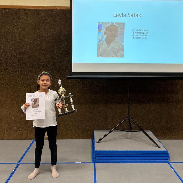 M&auml;dchen h&auml;lt eine Urkunde und einen Pokal vor einem Bildschirm mit ihrem Namen und Informationen.