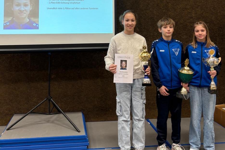 Drei junge Sportler stehen vor einer Pr&auml;sentation mit Informationen &uuml;ber Sofia Macovetchi und Pokalen.