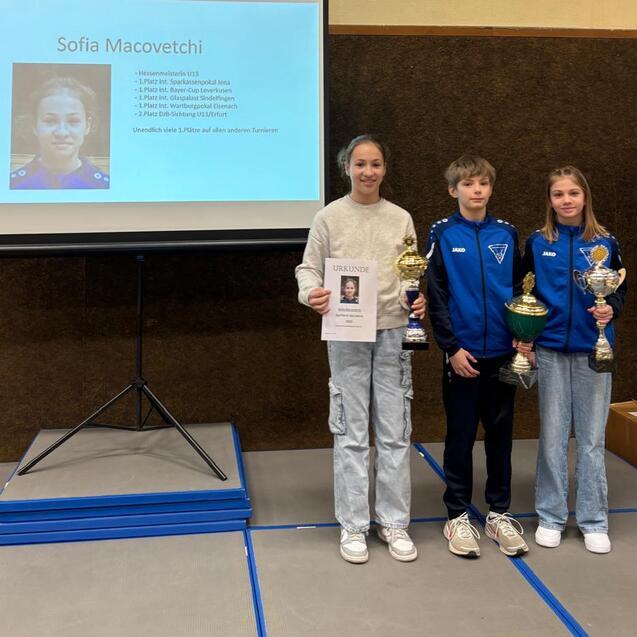 Drei junge Sportler stehen vor einer Pr&auml;sentation mit Informationen &uuml;ber Sofia Macovetchi und Pokalen.