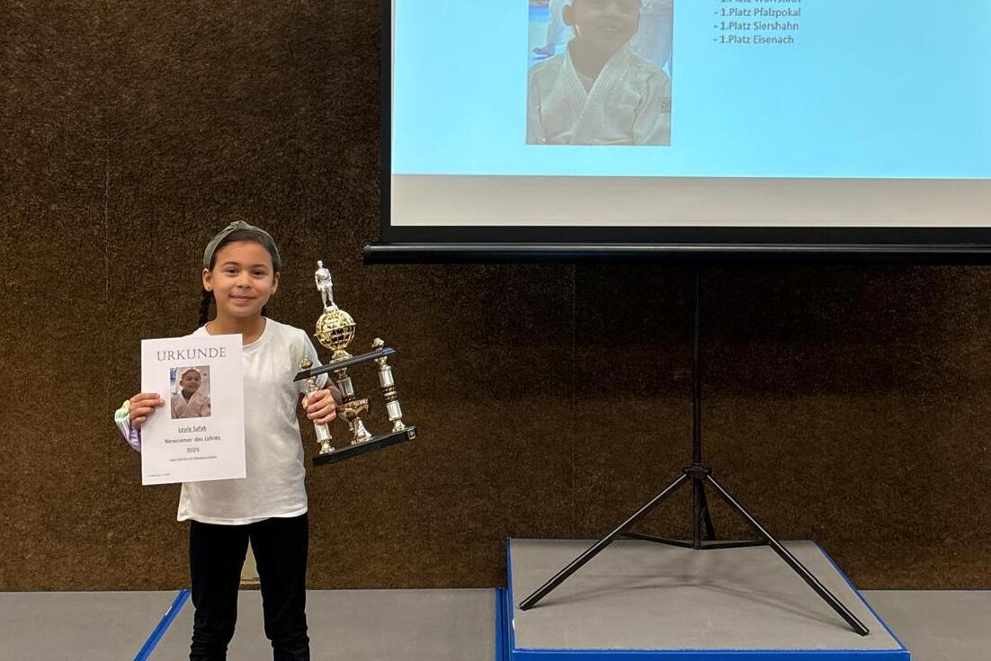 M&auml;dchen h&auml;lt eine Urkunde und einen Pokal vor einem Bildschirm mit ihrem Namen und Informationen.