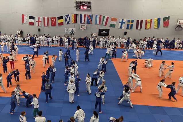 Gro&szlig;e Kampfsportszene mit vielen K&auml;mpfern auf blauen und orangefarbenen Matten in einer Halle, umgeben von Flaggen.