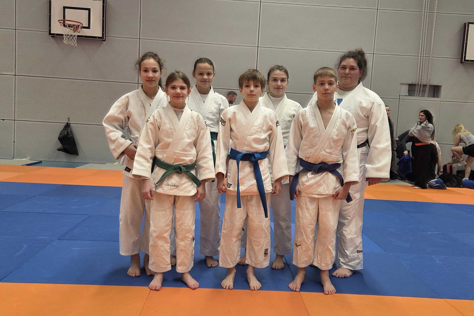 Gruppe von Judoka in weißen und blauen Anzügen auf einer Judomatte, posierend für ein Foto in einer Sporthalle.