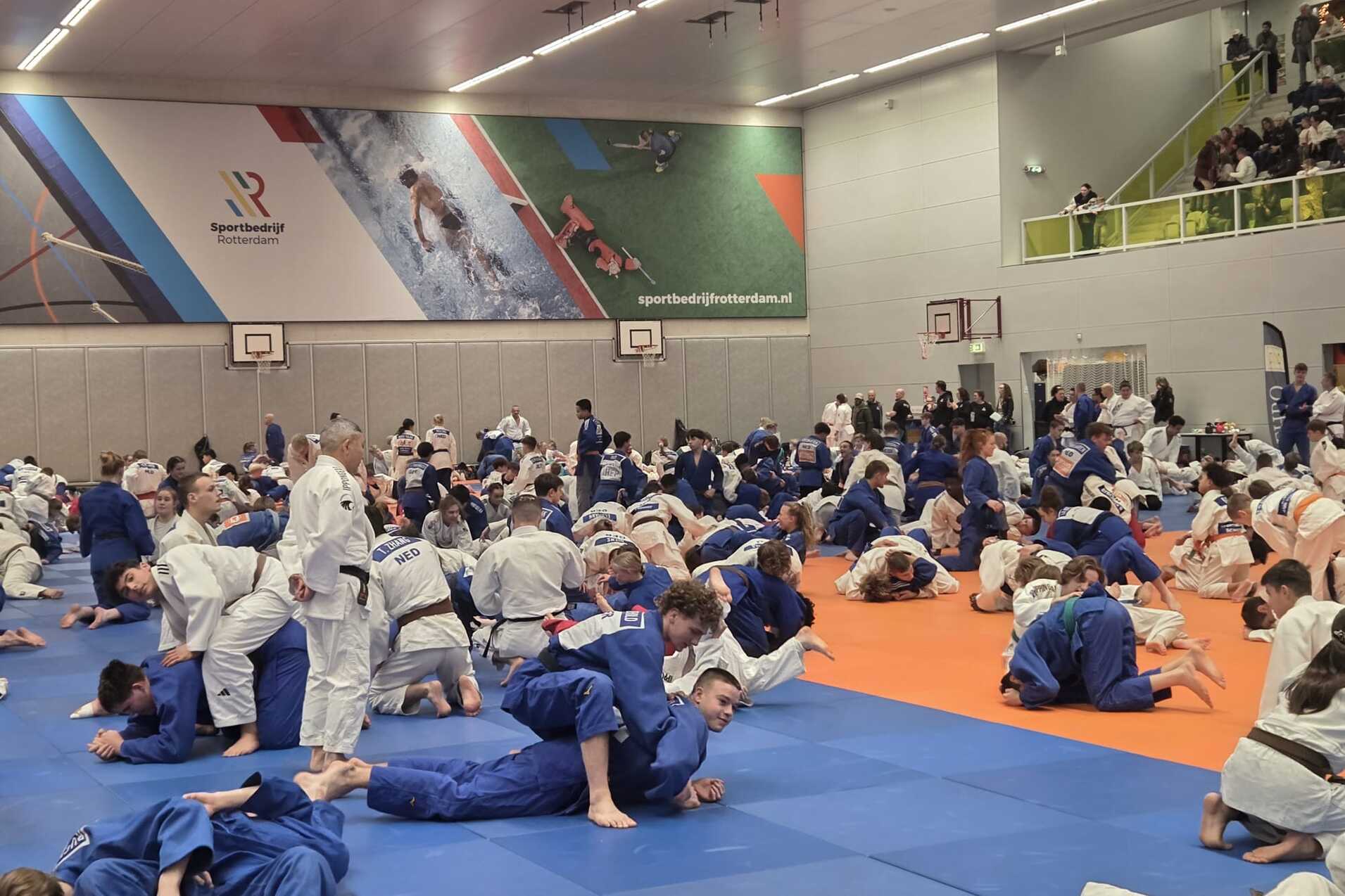 Vielbeiniger Judokreis mit Sportlern in weißen und blauen Judogis auf blauen und orangefarbenen Matten in einer Sporthalle.