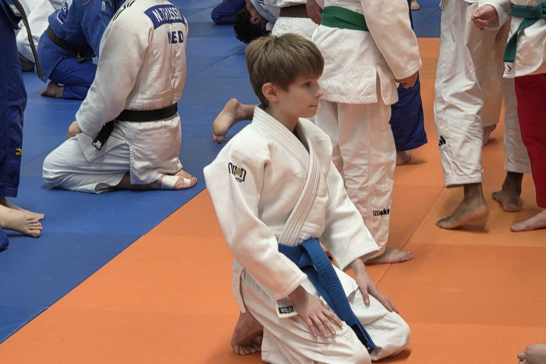 Ein Junge in einem weißen Judoanzug mit blauem Gürtel kniet auf einer orangefarbenen Matte, umgeben von anderen Judokas.