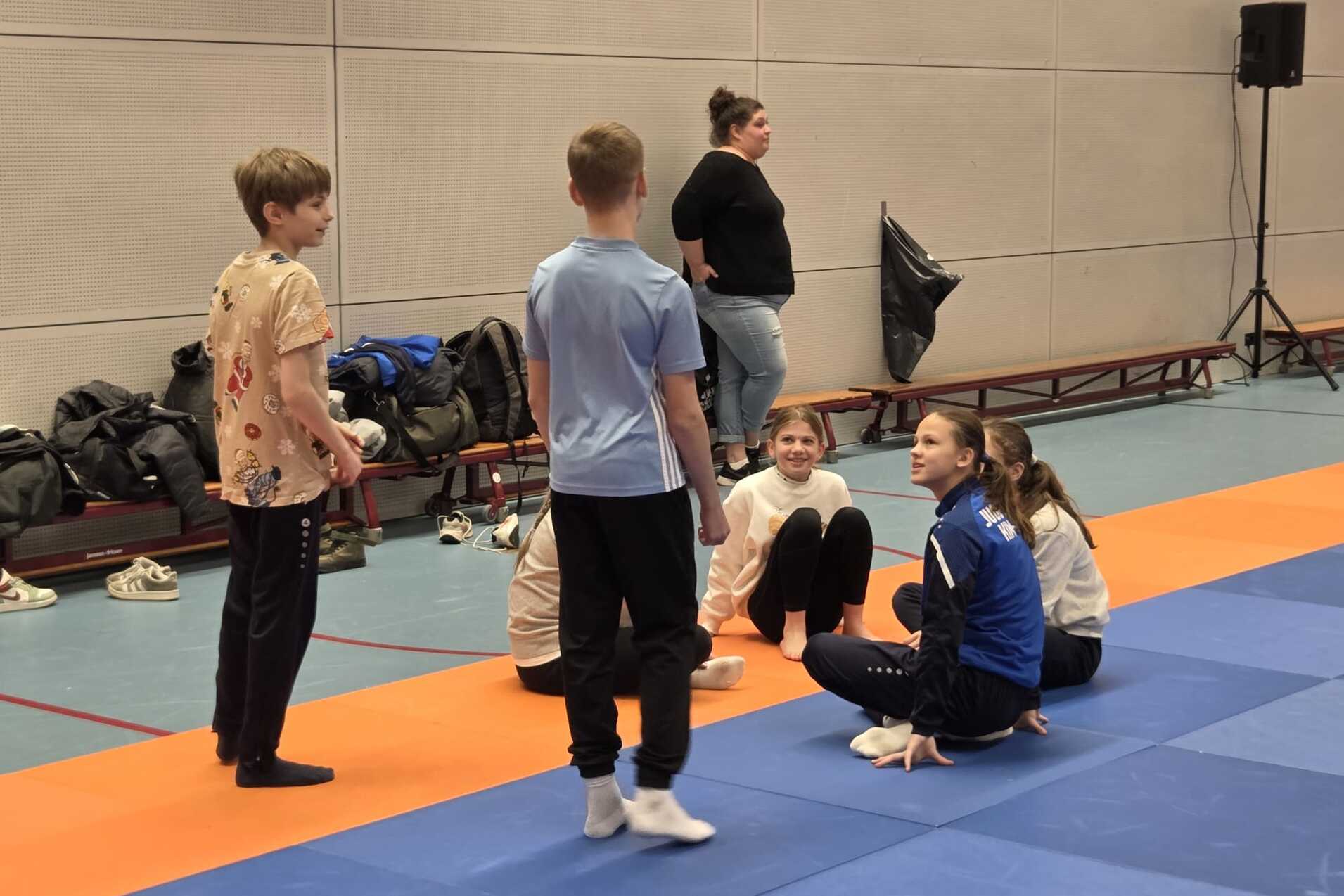 Sechs Kinder und ein Erwachsener stehen und sitzen in einer Sporthalle auf einer blauen Matte und orangefarbenem Bereich.