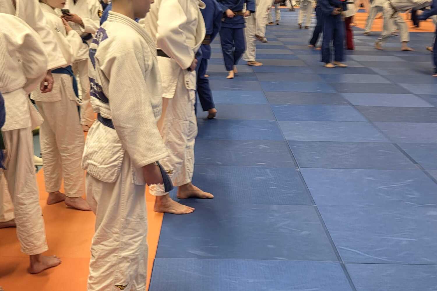 Gruppe von Judokas in weißen und blauen Gis, die auf blauen Matten in einer Halle stehen und sich auf das Training vorbereiten.