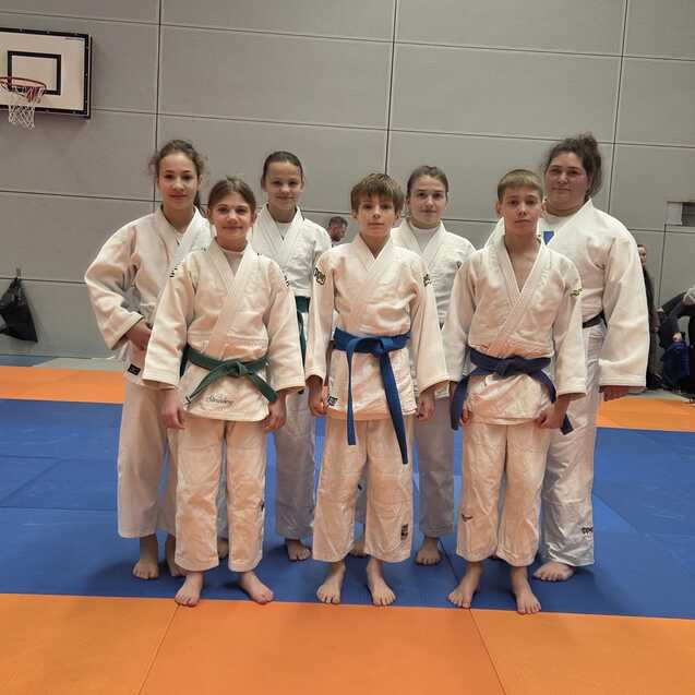 Gruppe von Judoka in wei&szlig;en und blauen Anz&uuml;gen auf einer Judomatte, posierend f&uuml;r ein Foto in einer Sporthalle.
