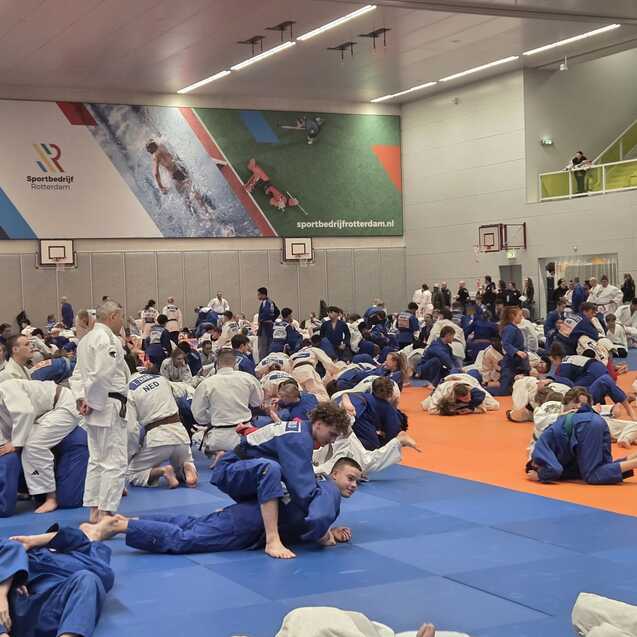 Vielbeiniger Judokreis mit Sportlern in wei&szlig;en und blauen Judogis auf blauen und orangefarbenen Matten in einer Sporthalle.