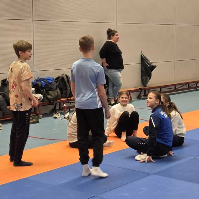 Sechs Kinder und ein Erwachsener stehen und sitzen in einer Sporthalle auf einer blauen Matte und orangefarbenem Bereich.