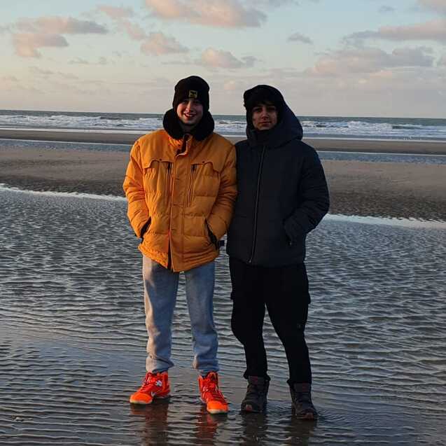 Zwei Personen stehen am Strand bei Sonnenaufgang auf feuchtem Sand vor dem Meer. Sie tragen warme Kleidung.