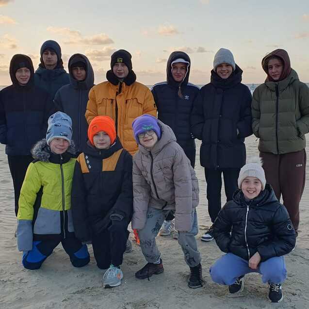 Gruppe von Kindern und Jugendlichen in warmen Winterjacken steht am Strand bei Sonnenuntergang.