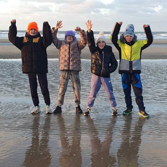 Vier Kinder in Winterkleidung stehen am Strand und winken vor einem ruhigen Meer bei bew&ouml;lktem Himmel.