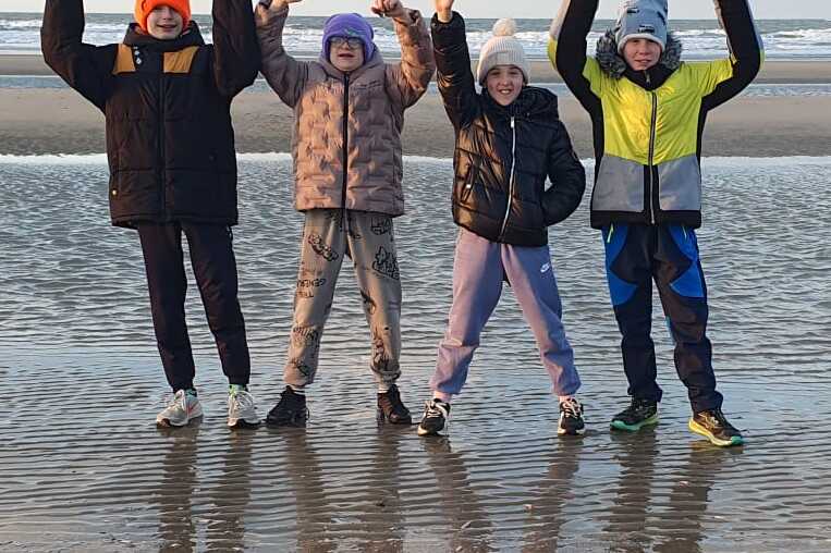 Vier Kinder in Winterkleidung stehen am Strand und winken vor einem ruhigen Meer bei bewölktem Himmel.