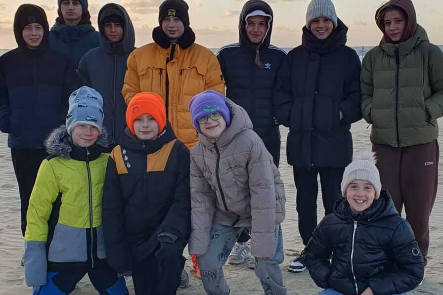 Gruppe von Kindern und Jugendlichen in warmen Winterjacken steht am Strand bei Sonnenuntergang.