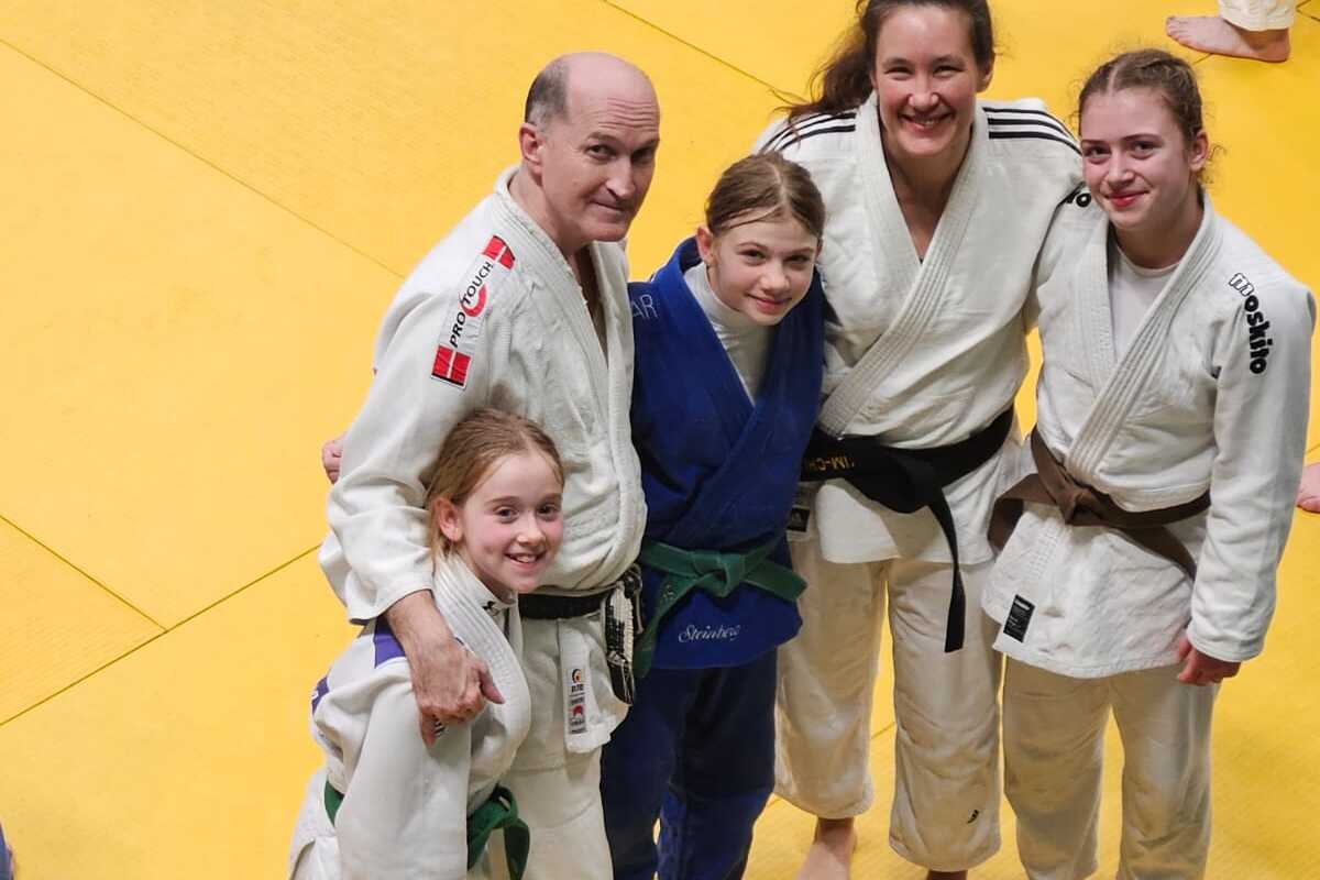 Fünf Personen in Judogis posieren lächelnd auf einer gelben Judomatte.