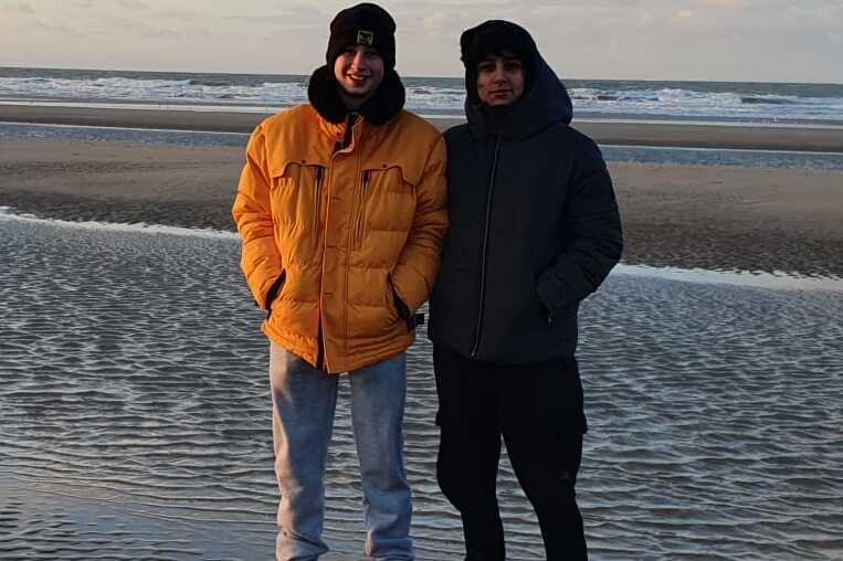 Zwei Personen stehen am Strand bei Sonnenaufgang auf feuchtem Sand vor dem Meer. Sie tragen warme Kleidung.