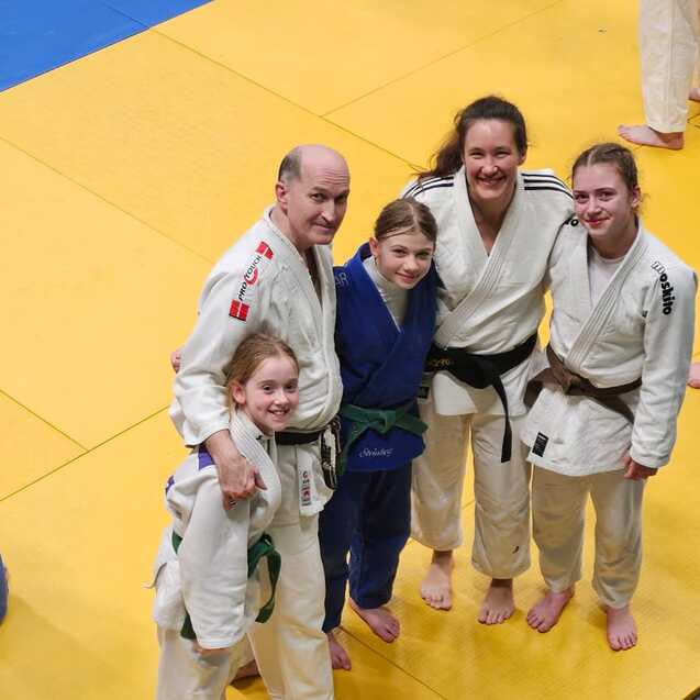 F&uuml;nf Personen in Judogis posieren l&auml;chelnd auf einer gelben Judomatte.