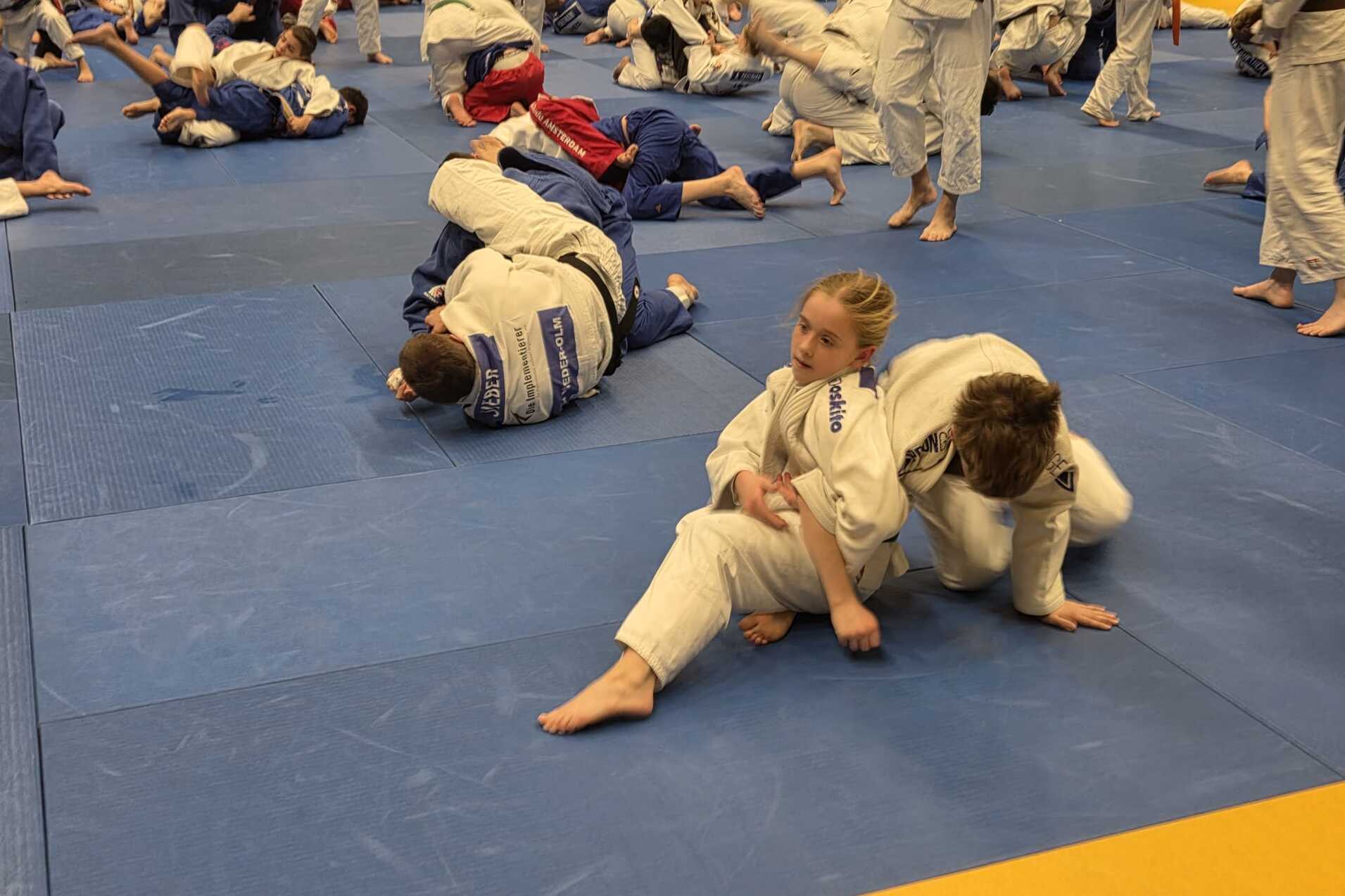 Eine Gruppe von Judokas in weißen und blauen Anzügen trainiert auf einer blauen Matte in einem Sportzentrum.