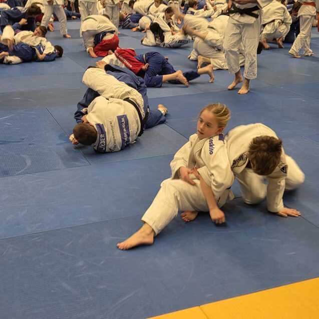 Eine Gruppe von Judokas in wei&szlig;en und blauen Anz&uuml;gen trainiert auf einer blauen Matte in einem Sportzentrum.