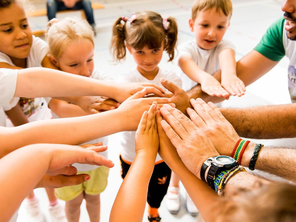 Kinder und Erwachsene legen ihre Hände in einem Kreis zusammen, symbolisieren Zusammenarbeit und Gemeinschaft.