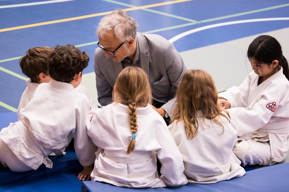 Ein Trainer spricht mit einer Gruppe von fünf Kindern in Judoanzügen auf einer Sportmatte.