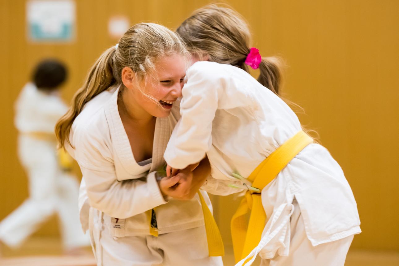 Zwei Mädchen im Judoanzug lachen fröhlich miteinander auf einer Judomatte. Eines trägt einen gelben Gürtel.
