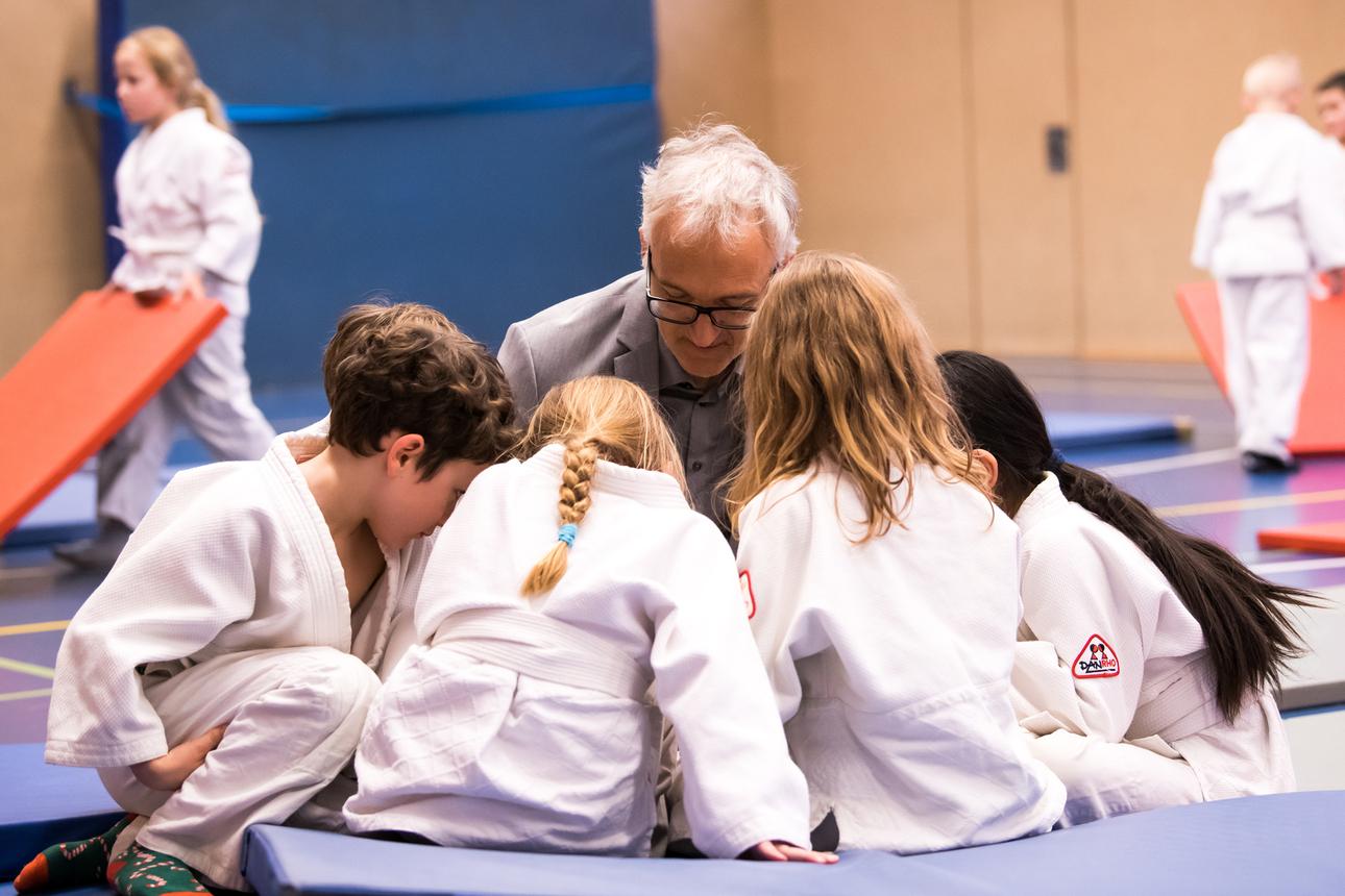 Sechs Kinder in Judogis beugen sich um einen älteren Trainer in einem Sportraum, während andere Kinder im Hintergrund spielen.
