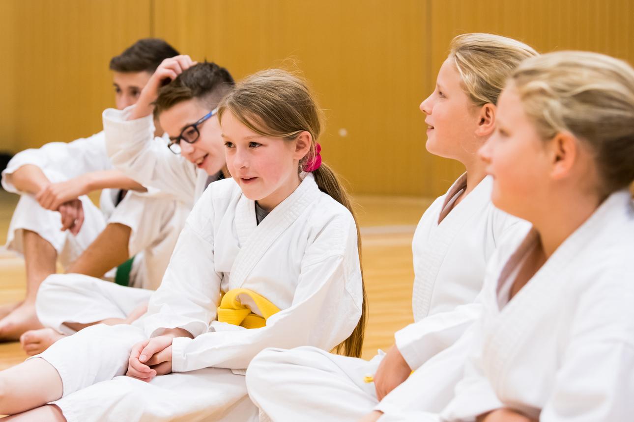 Fünf Kinder in weißen Karateanzügen sitzen in einer Trainingshalle und unterhalten sich.