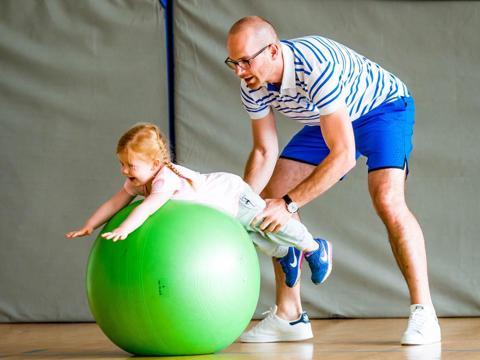 Ein Erwachsener hilft einem Kind, auf einem großen grünen Gymnastikball zu balancieren, in einer Sporthalle.