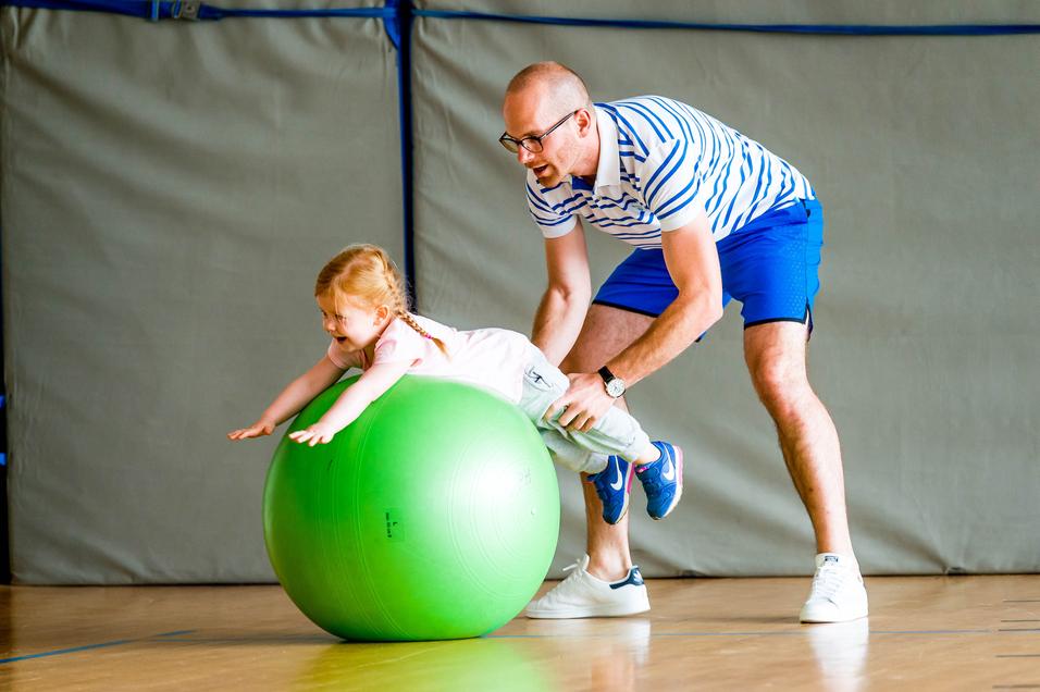 Ein Erwachsener hilft einem Kind, auf einem großen grünen Gymnastikball zu balancieren, in einer Sporthalle.